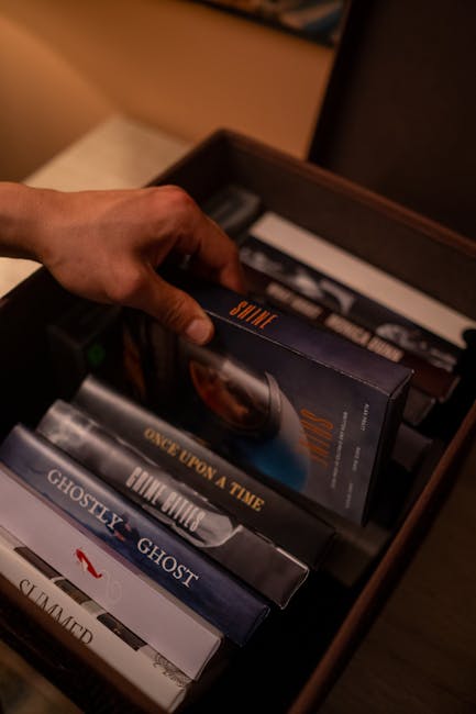 A hand selecting a VHS tape from a box of various nostalgic movie covers indoors.